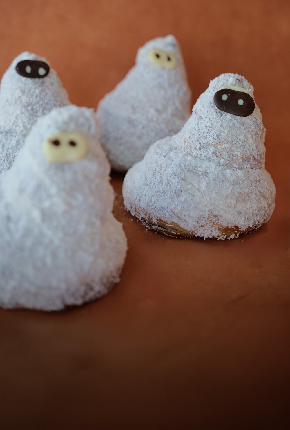 Four ghost-shaped pastries coated in white coconut flakes against an orange background.